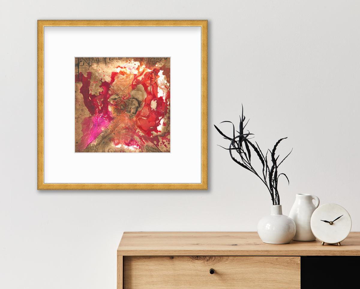 A gold, red and pink abstract print in a gold frame with a mat hangs on a white wall above a wood cabinet with a white modern clock, a small white pitcher and a white vase with dark green stems.