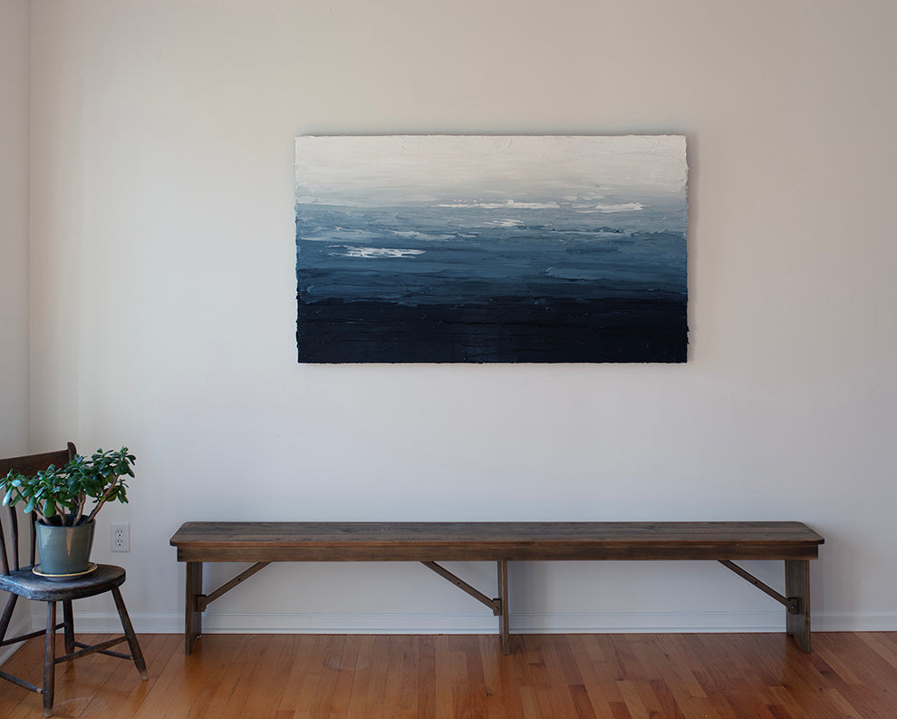 A blue, white and navy very textured abstract painting hangs on a white wall above a bench in the artists studio..
