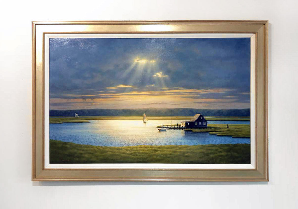 A painting of a bay house with sailboats, framed in a gold frame with a linen lining. On a white wall.
