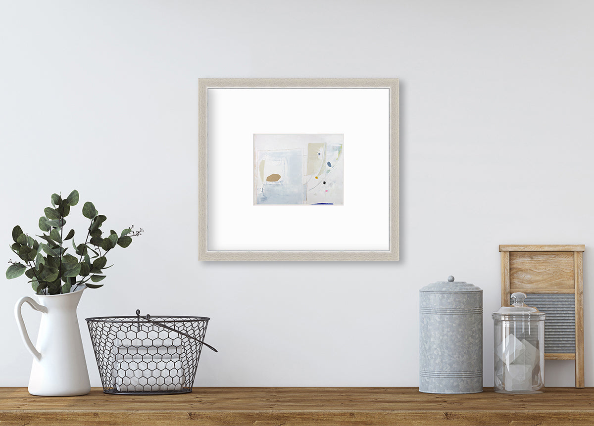 A white, blue, beige and light green abstract print in a silver frame with a mat hangs on a white wall above a wood surface with decorative accessories.
