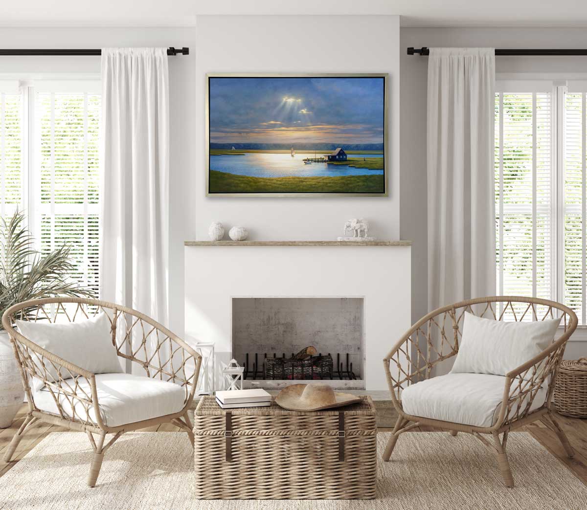 A realist coastal print in a silver floater frame hangs on a white wall above a fireplace mantle, two woven chairs with white cushions and a woven trunk.