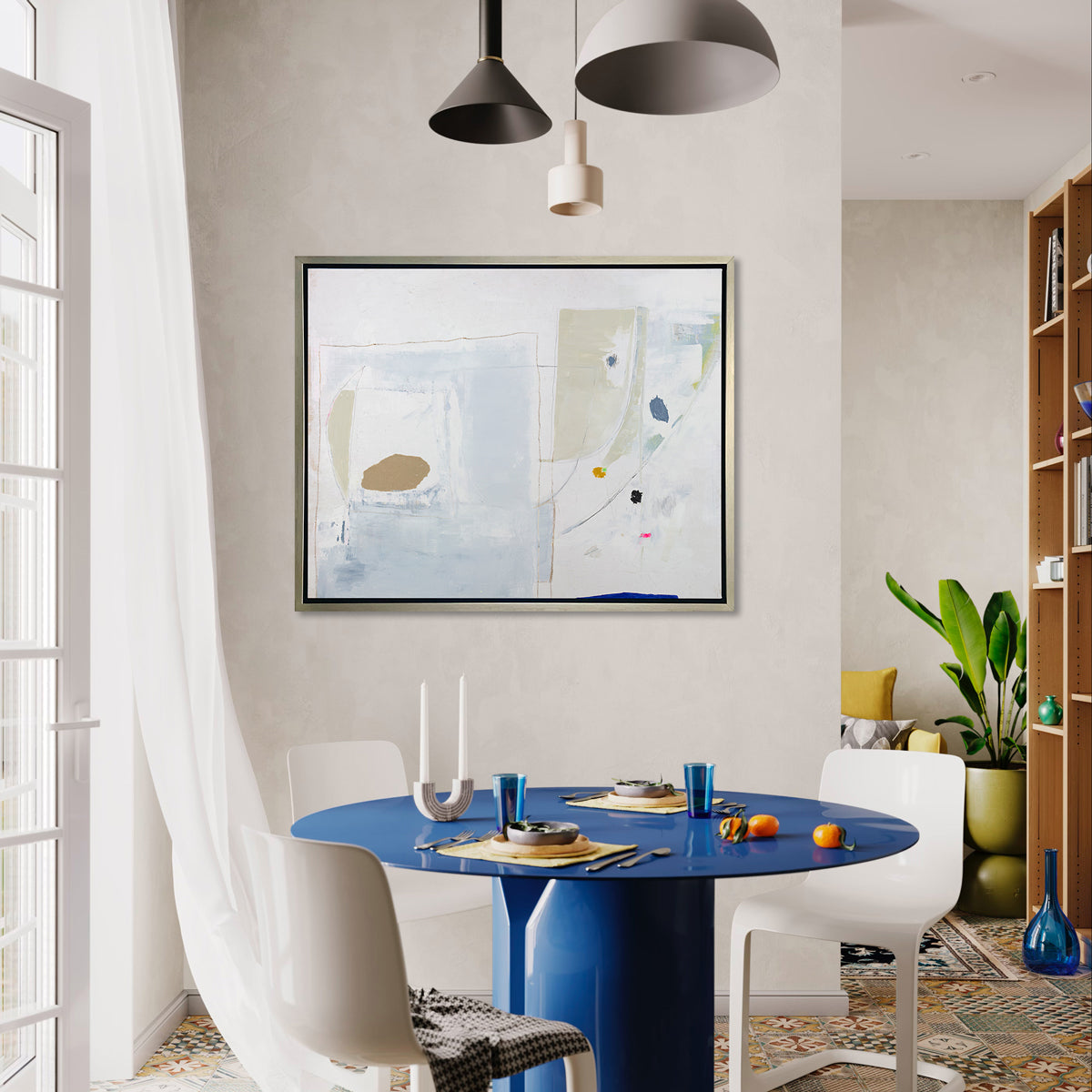 A white, blue, beige and light green abstract print in a silver floater frame hangs on a white wall above a modern blue pedestal table with place settings, three white chairs surrounding and a tall bookshelf in the background.