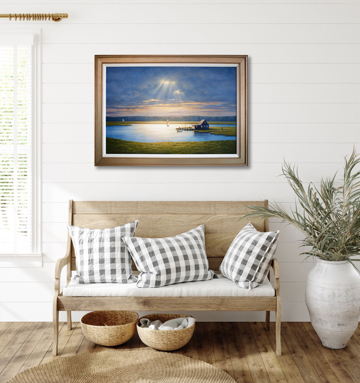 A landscape painting in a gold frame hangs above a wooden seat on a white wall, next to a large potted plant and a window on the other side. 