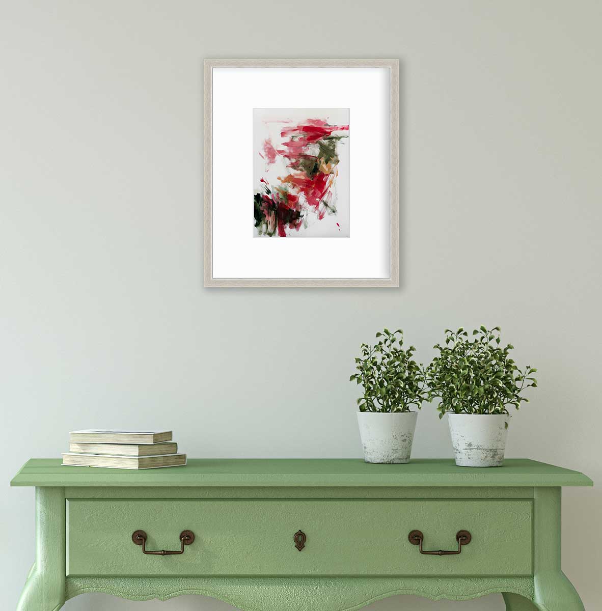 A red, green and white abstract print in a silver frame with a mat hangs on a white wall above a green console table with a stack of books and two potted plants.