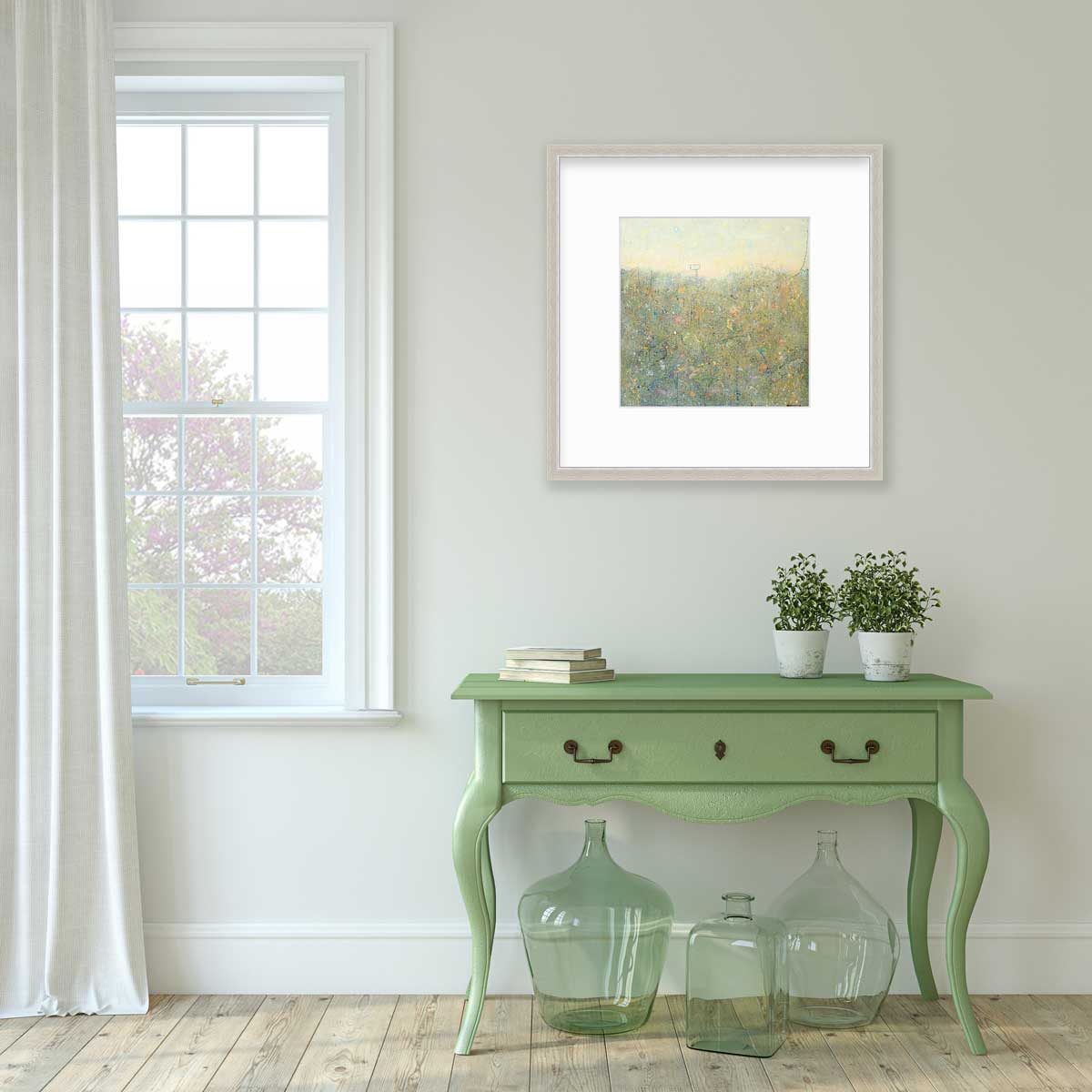 A yellow-green abstract landscape in a silver frame with a mat hangs on a white wall above a green console table with a stack of books, two potted plants and three green glass bottles.