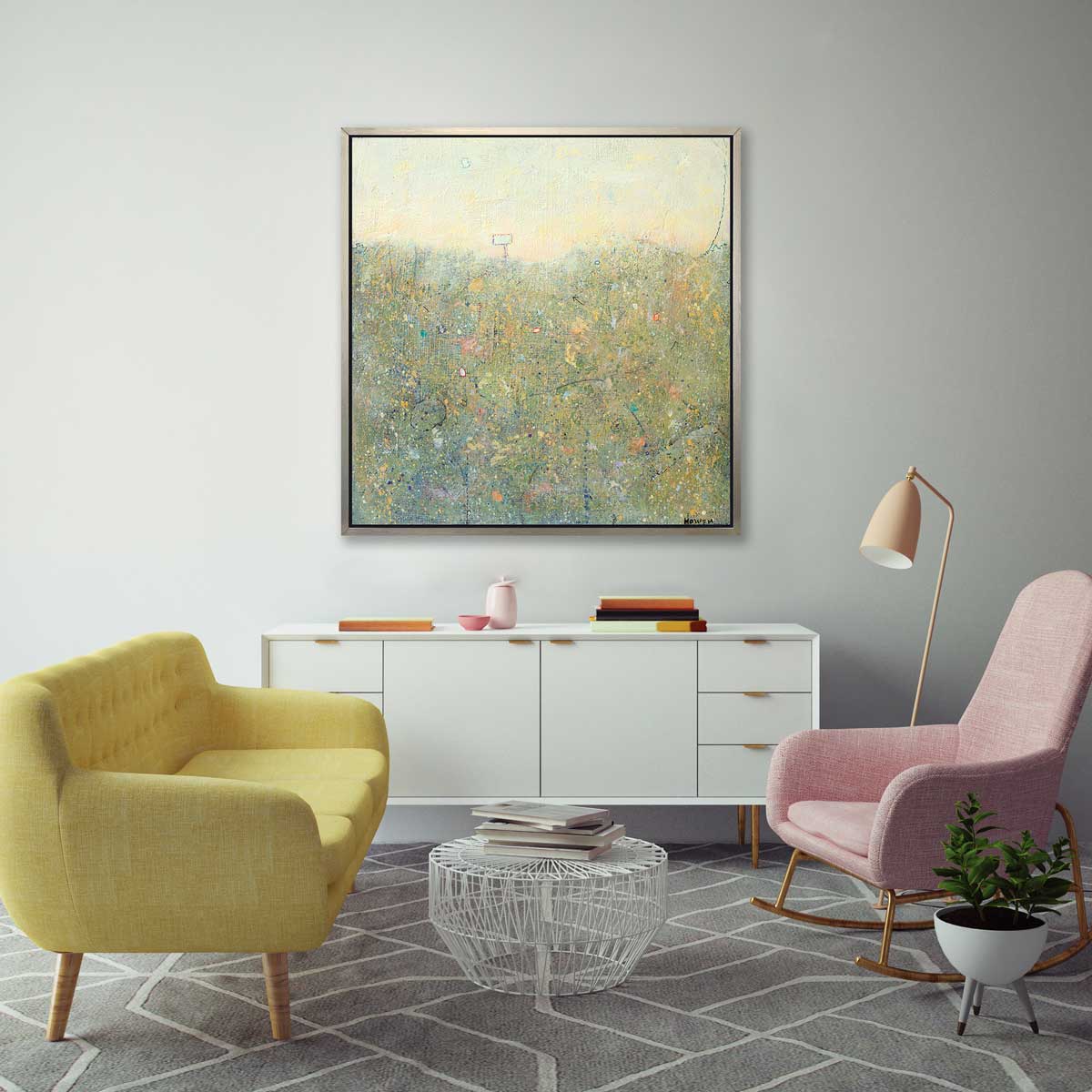 A green and light yellow abstract landscape print in a silver floater frame hangs on a grey wall above a white sideboard with books, a yellow tufted sofa and a pink rocking chair.