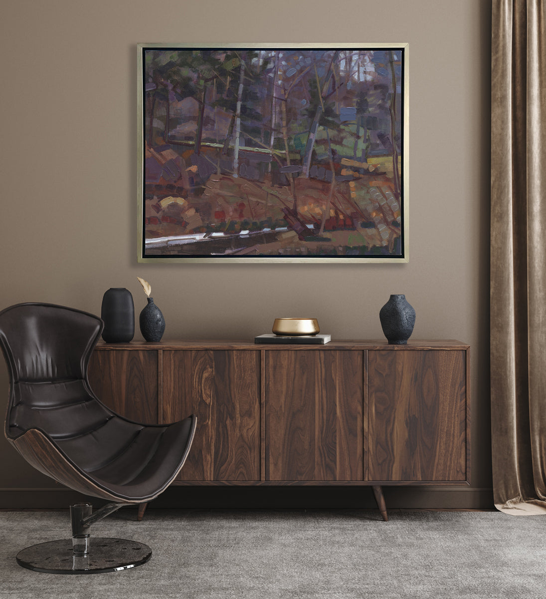 A brown, grey and green abstract landscape print in a silver floater frame hangs on a beige wall above a dark wood sideboard with black vases, a gold decorative dish and a brown modern chair.