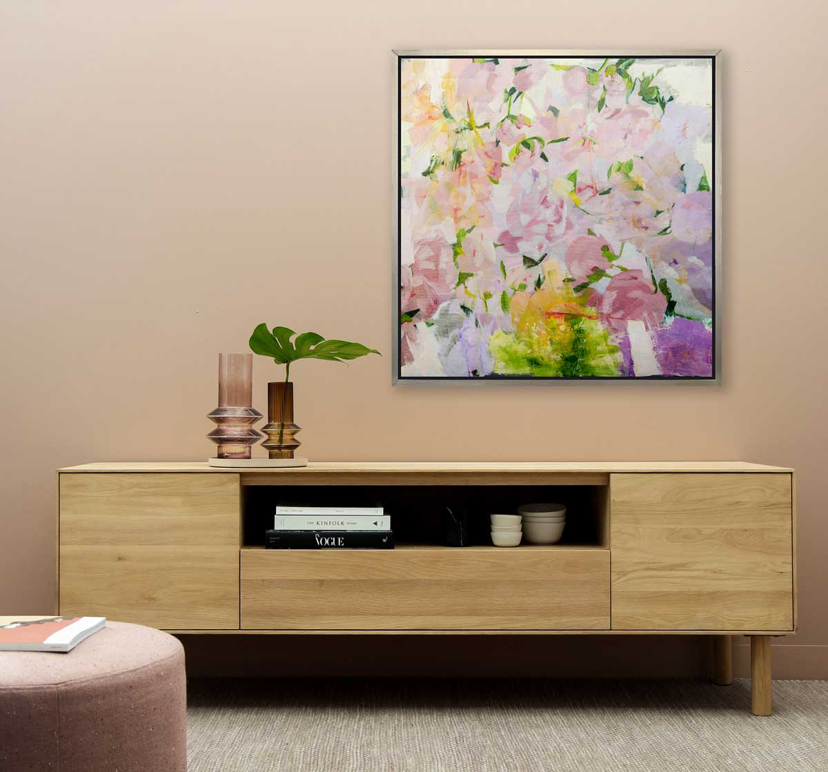 A multicolored floral abstract print hangs on a beige wall above a wood console cabinet with books, dishes and two opaque glass vases with a giant leaf stem.