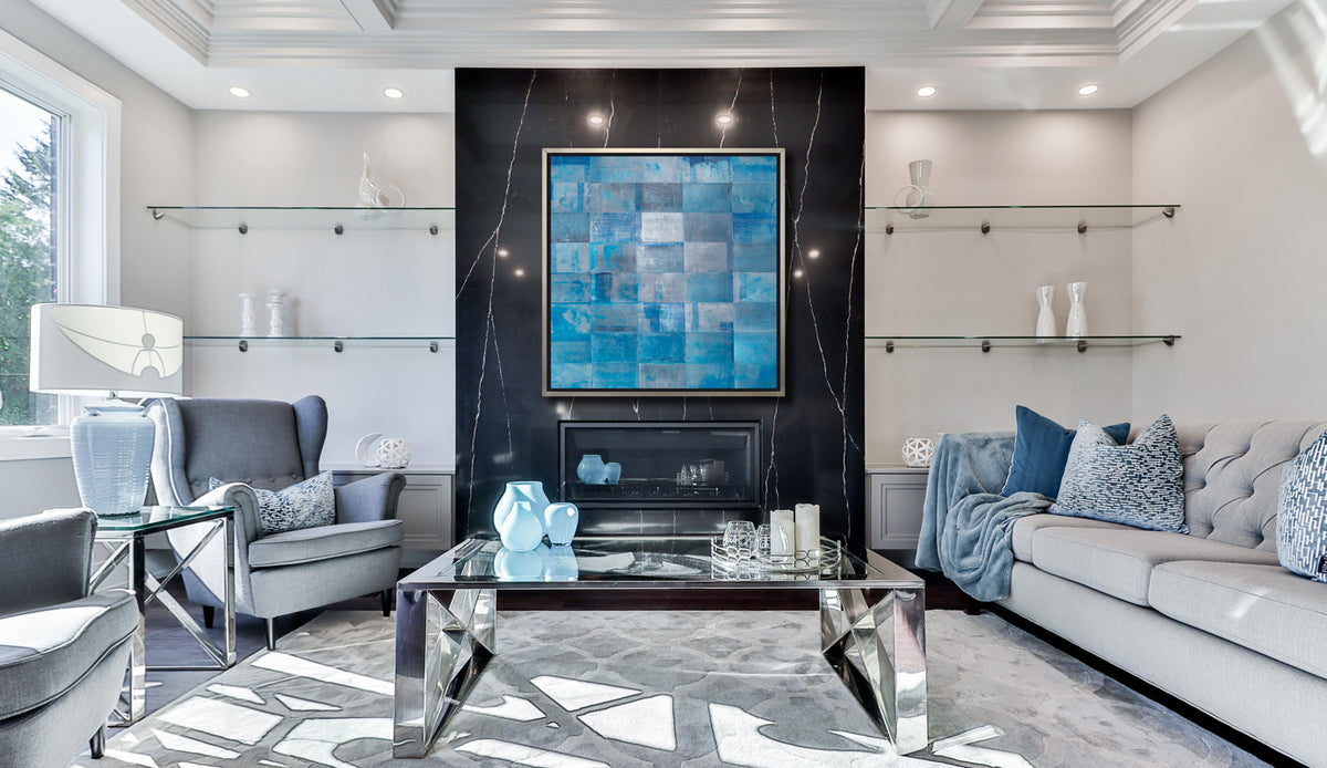 A blue geometric print in a silver floater frame hangs on a black marble wall above a fireplace, a light grey sofa with blue pillows and blankets, a grey high back chair and a glass coffee table with blue vases.