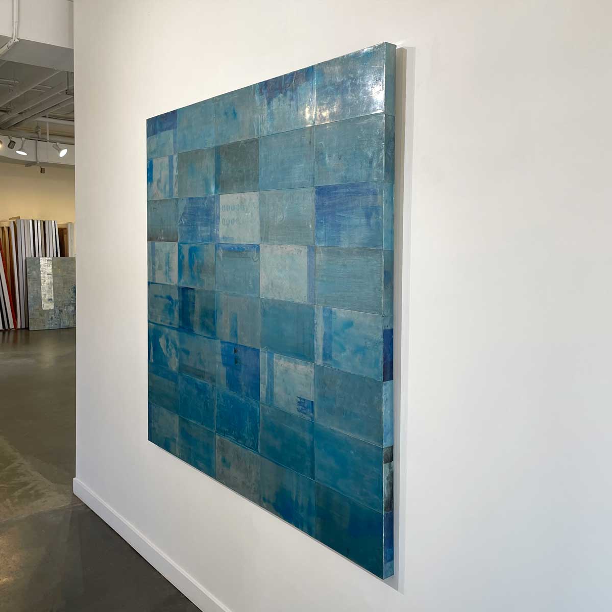 An abstract painting with a grid appearance and a blue wash by Ned Martin hangs on a white wall with a view from the right. 