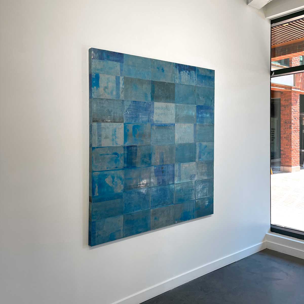 An abstract painting with a grid appearance and a blue wash by Ned Martin hangs on a white wall with a view from the left. 