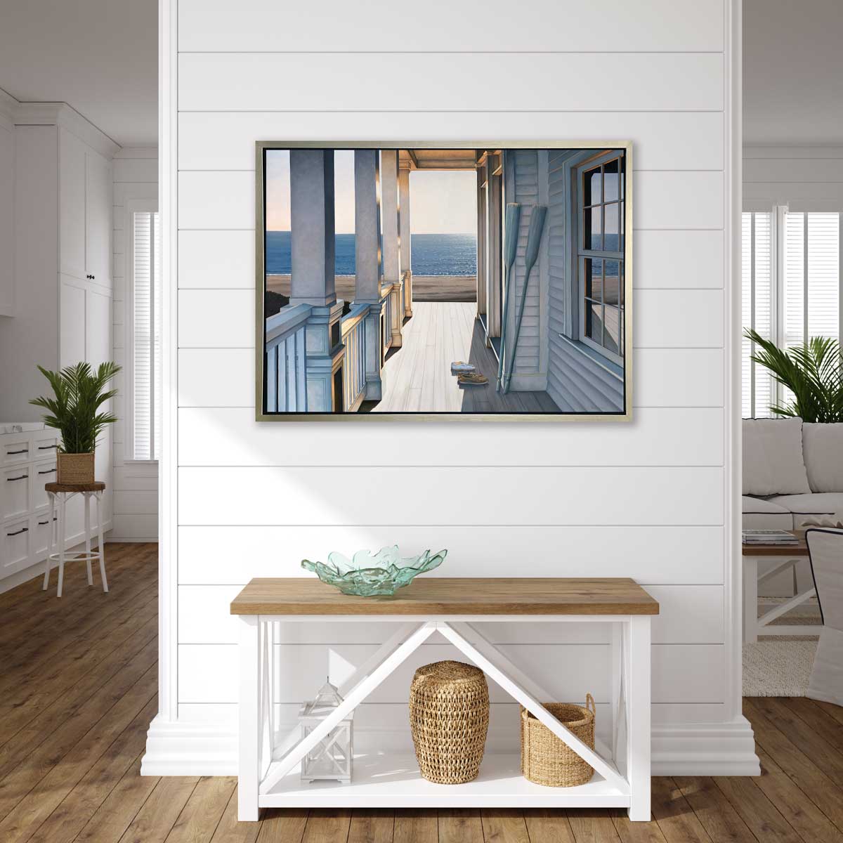 A print of a sunlit porch overlooking the water with two oars and two pairs of shoes on the deck in a silver floater frame hangs on a white shiplap wall above a wood and white console table with a teal decorative glass bowl, a white lantern and two woven baskets.