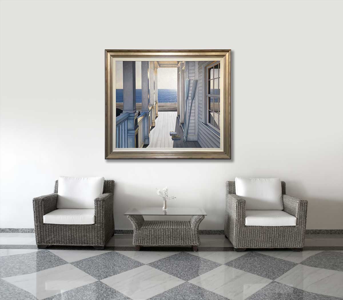 A realistic seascape painting in a warm silver frame hangs on a white wall above two matching dark woven chairs with white cushions and a coordinating coffee table with a glass top.
