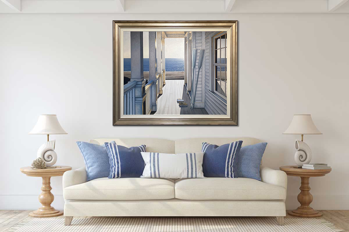 A realistic seascape painting in a warm silver frame hangs on a white wall above an ivory sofa with blue pillows and two wooden end tables with matching seashell table lamps.