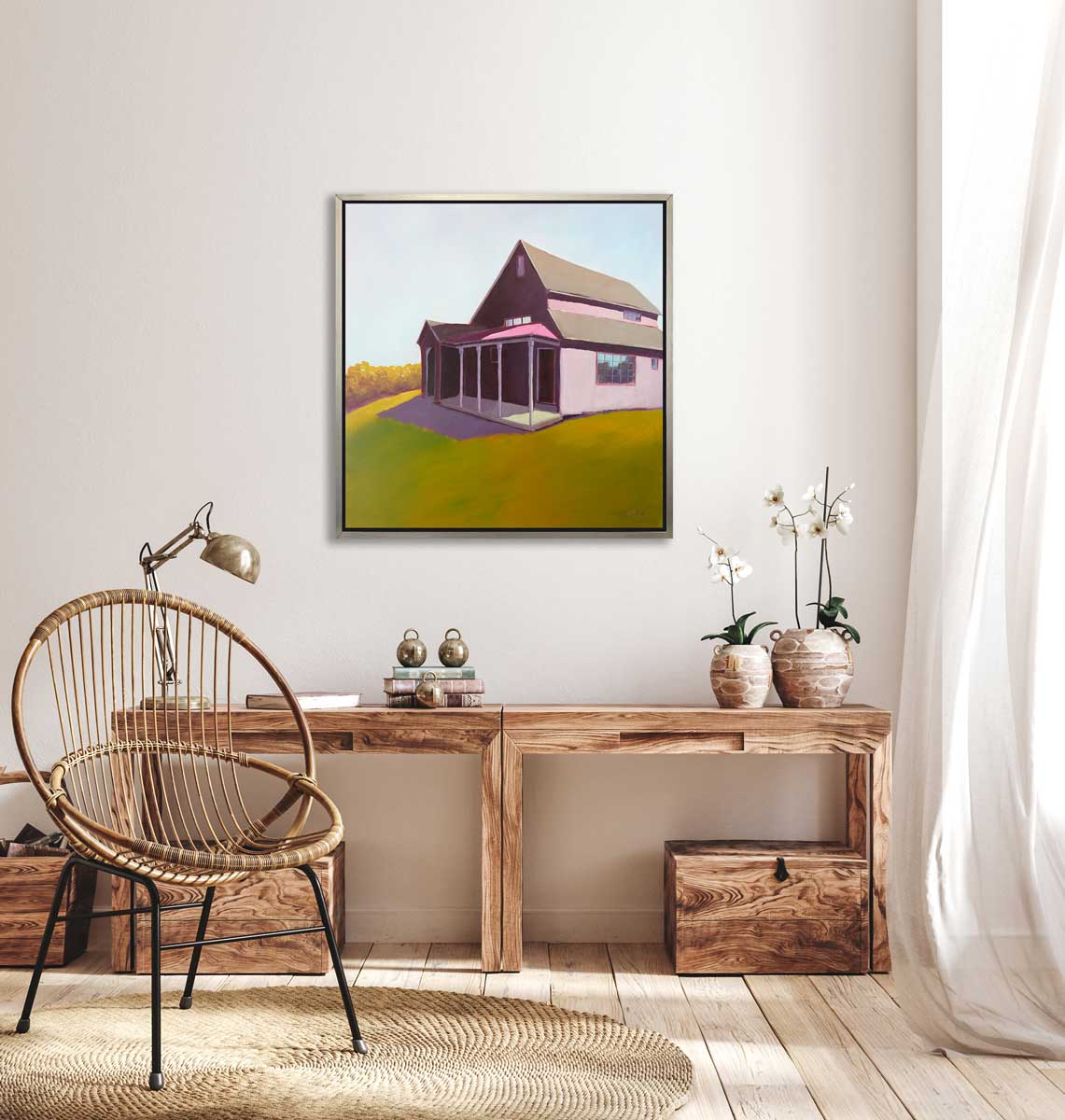 A raspberry, chartruse and light blue contemporary house print in a silver floater frame hangs on a white wall above a natural wood desk with a stack of books, a chrome desk lamp, two potted white orchids and a woven chair.