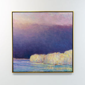 A painting of yellow trees on a lakeside with a purple and pink sky with the sun peaking in from upper left hand corner in a gold floater frame hangs on a white wall in the gallery.