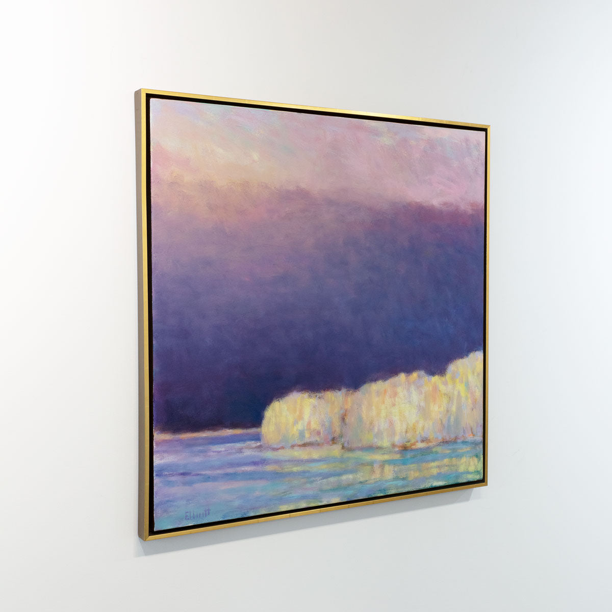A painting of yellow trees on a lakeside with a purple and pink sky with the sun peaking in from upper left hand corner in a gold floater frame hangs on a white wall in the gallery, viewed from the left.