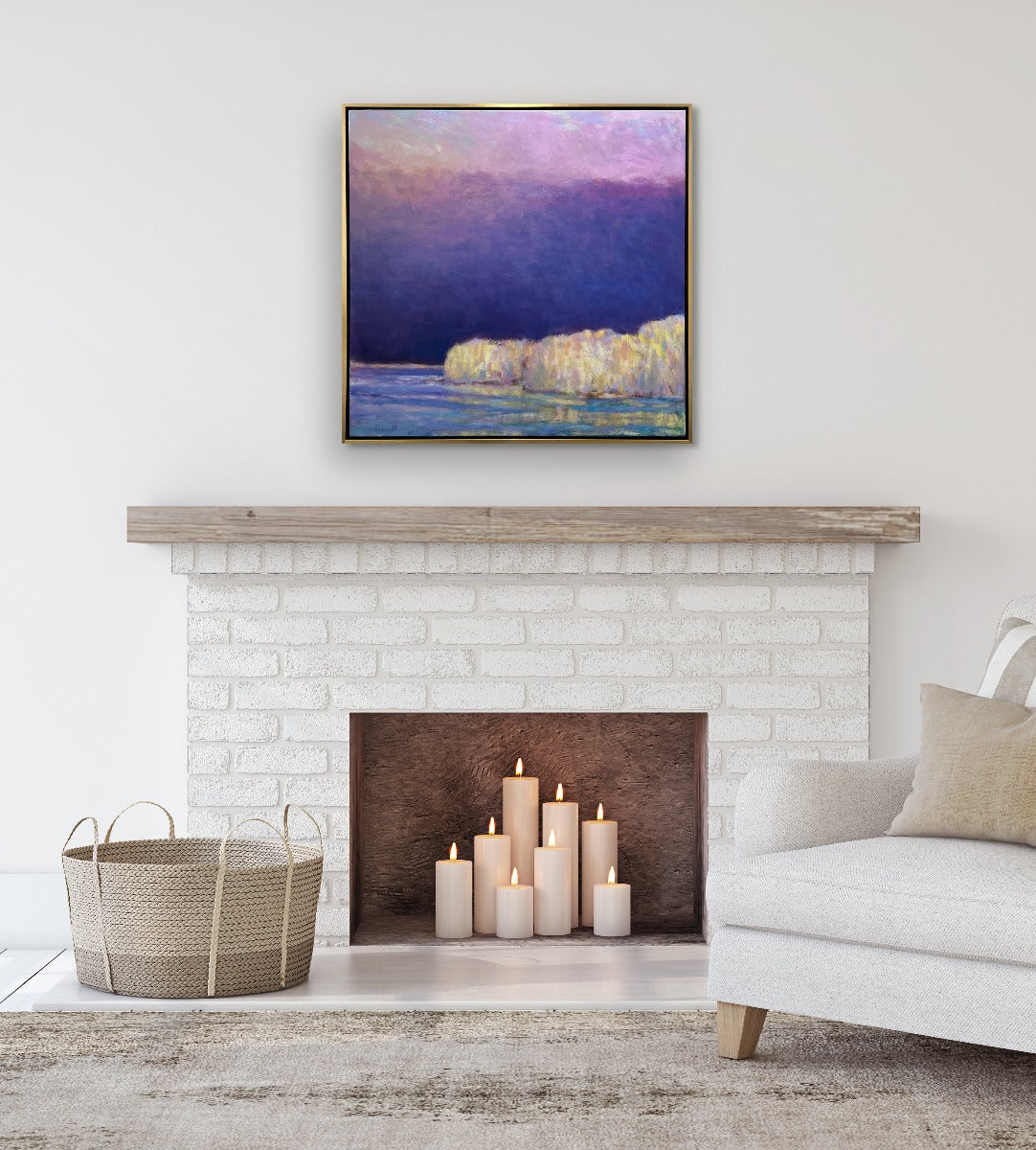A painting of yellow trees on a lakeside with a purple and pink sky with the sun peaking in from upper left hand corner by Ken Elliott in a gold floater frame hangs on a white wall above a natural wood mantle and white brick fireplace. The fireplace holds different sized lite candles and a light beige arm chair to the right. 