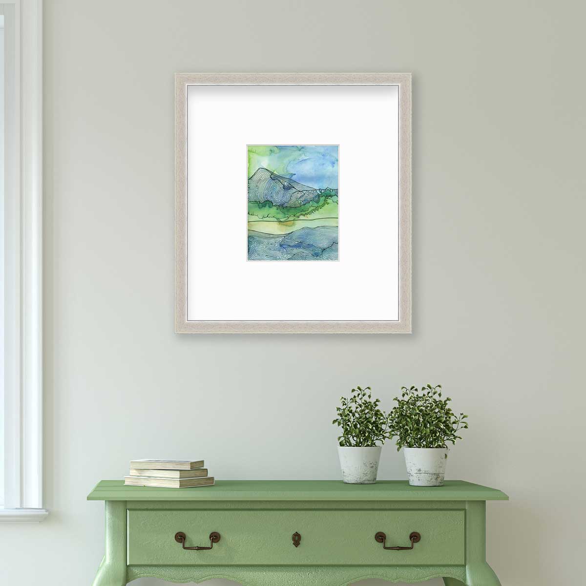 A blue and green landscape illustration print framed in a silver frame with a mat hangs on a light grey wall above a green console table with a stack of books and two potted plants.