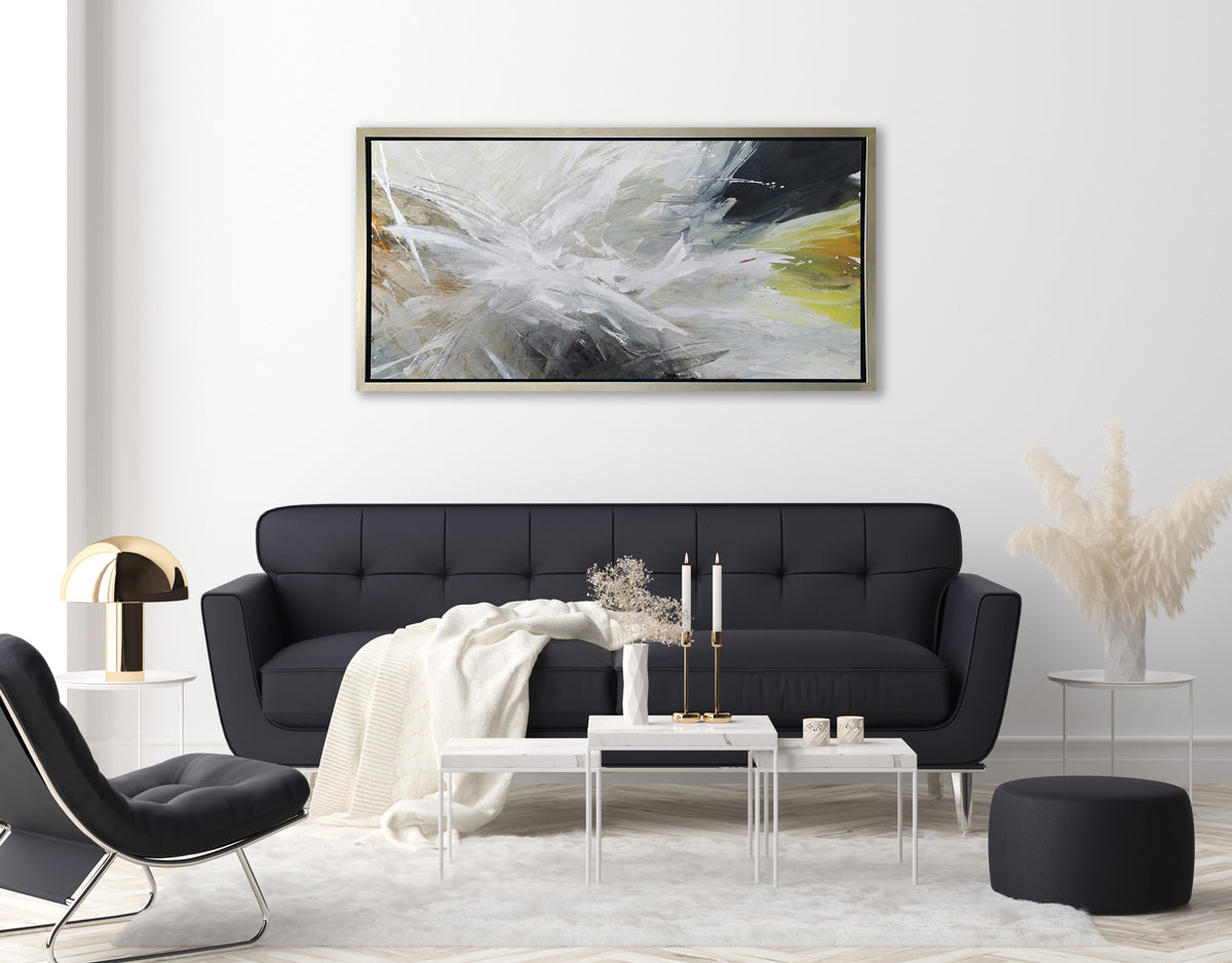 A white, black, grey and gold abstract print in a silver floater frame hangs on a white wall above a black tufted sofa with a white throw blanket, a modern black chair and a white two-tiered elongated coffee table.