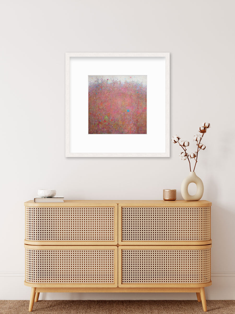 A red abstract landscape print in a whitewashed frame with a mat hangs on a white wall above a wood console cabinet with a book, a white bowl, a copper candleholder and a white modern vase with cotton stems.