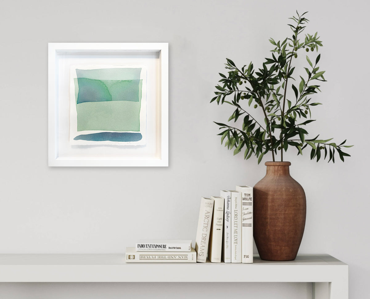 Blue and green shapes overlap in this painting by Nealy Hauschildt hangs in a white frame floating above a white matte on a white wall above a white surface with a stack of books and a greenery filled terracotta vase.