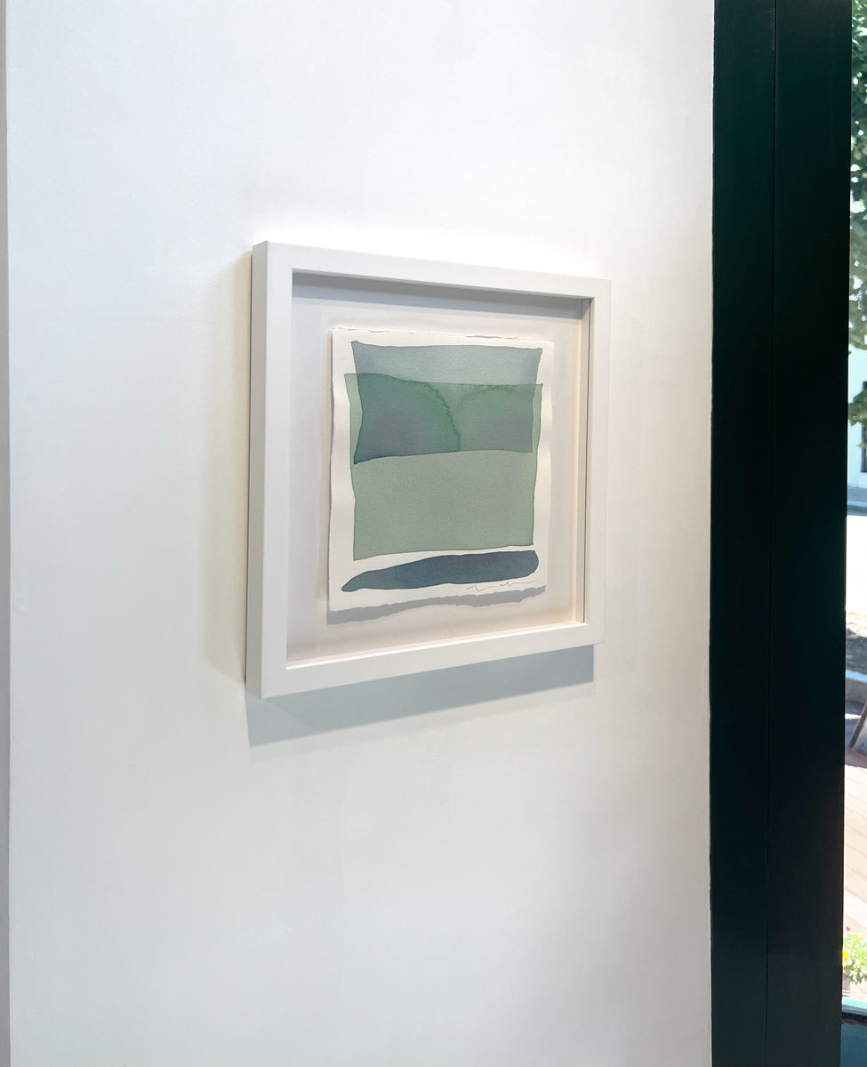 A left side-angle of blue and green shapes overlap in this painting by Nealy Hauschildt in a white frame floating above a white matte hangs on a white wall.