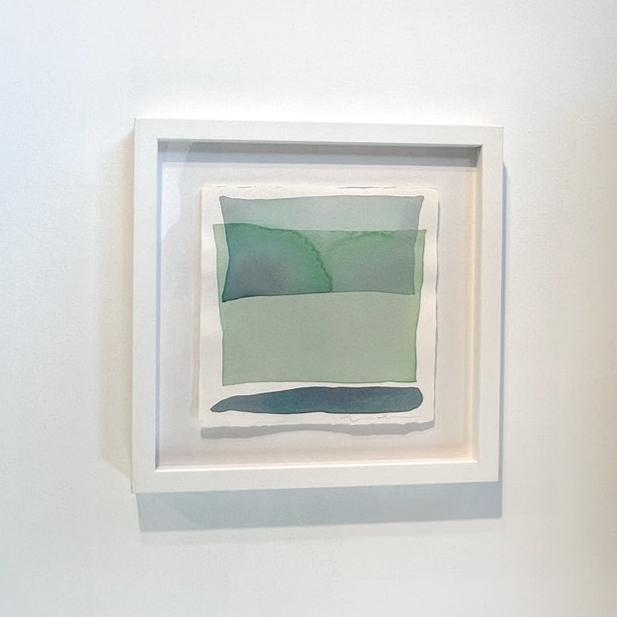 Blue and green shapes overlap in this painting by Nealy Hauschildt in a white frame floating above a white matte hangs on a white wall.