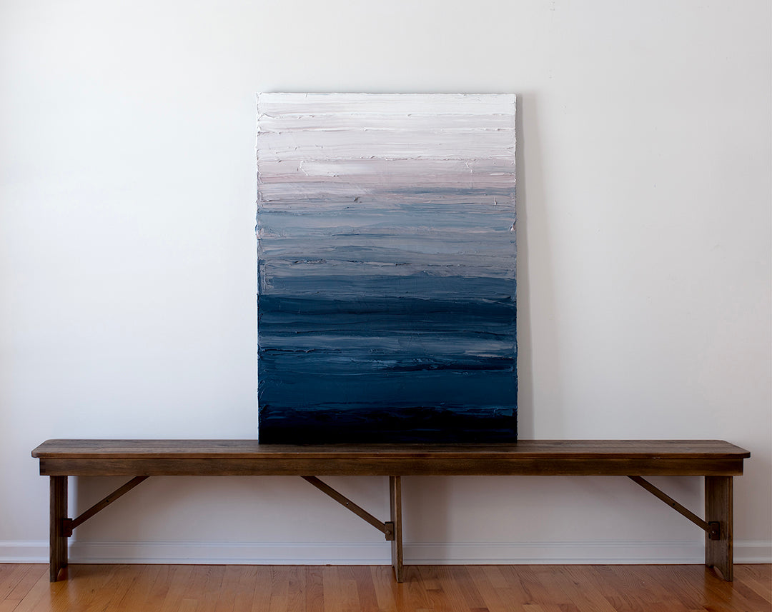A tonal blue, light pink and white textured abstract painting sits on a wood bench leaning on white wall.