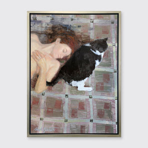 A print of a red-haired woman lays on a red and grey checkered floor next to a black and white cat in a silver floater frame hangs on a white wall.