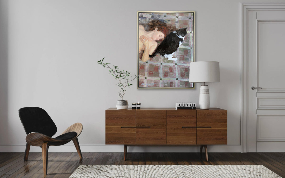 A print of a red-haired woman lays on a red and grey checkered floor next to a black and white cat in a silver floater frame hangs on a white wall above a dark wood credenza with a white table lamp, a stack of books, two small votives, a small potted plant and a modern black and wood chair.