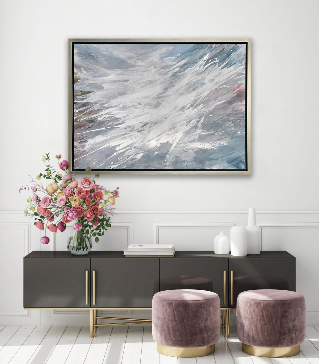 A grey, white and blue abstract print in a silver floater frame hangs on a white wall above a black credenza with an arrangement of white vases, a stack of books, a large floral arrangement and two mauve ottomans on the floor.