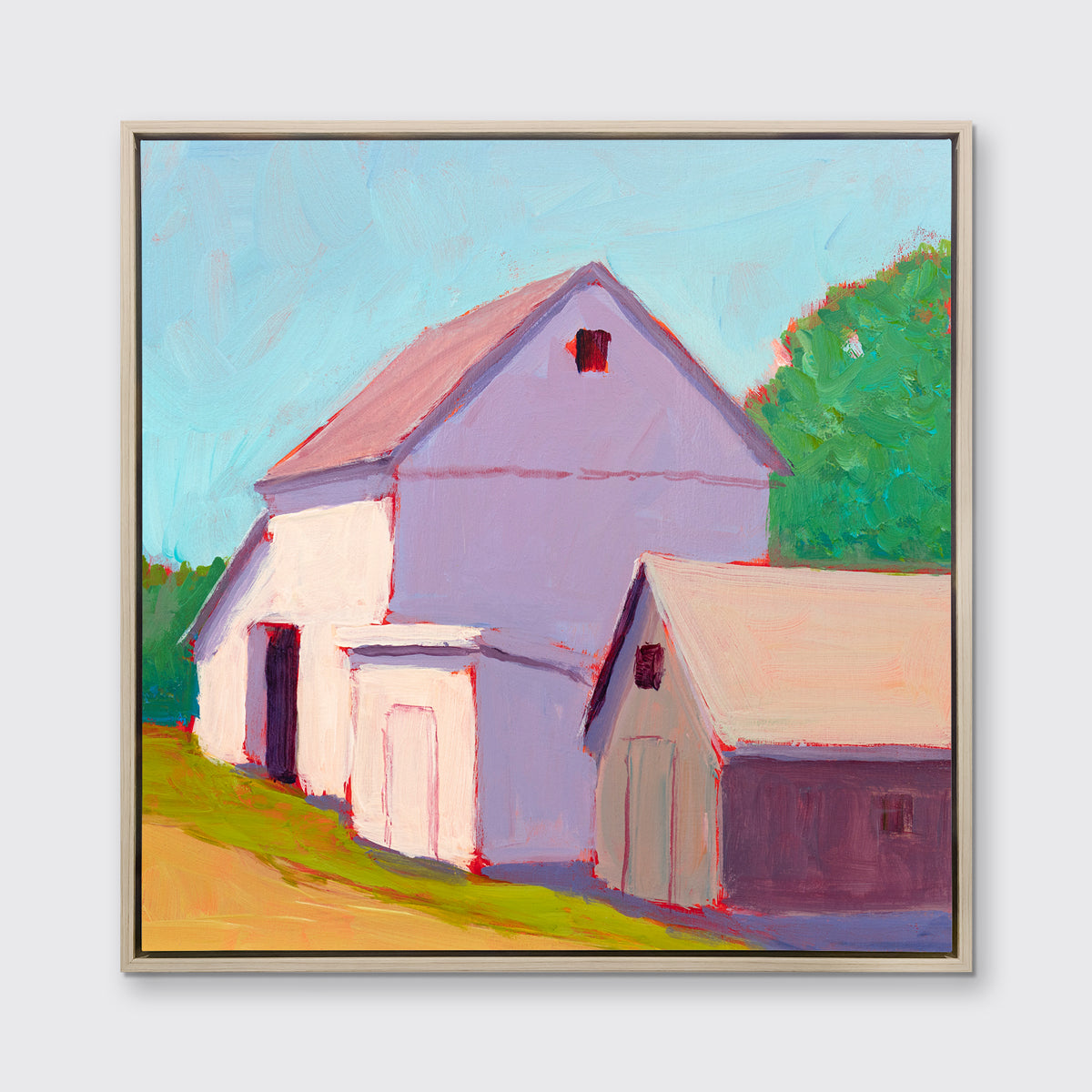 A print of a ivory barn, a smaller tan barn, a blue sky, green trees and a tan road in a white oak wood floater frame hangs on a white wall.