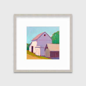 A print of a ivory barn, a smaller tan barn, a blue sky, green trees and a tan road in a silver frame with a mat hangs on a white wall.