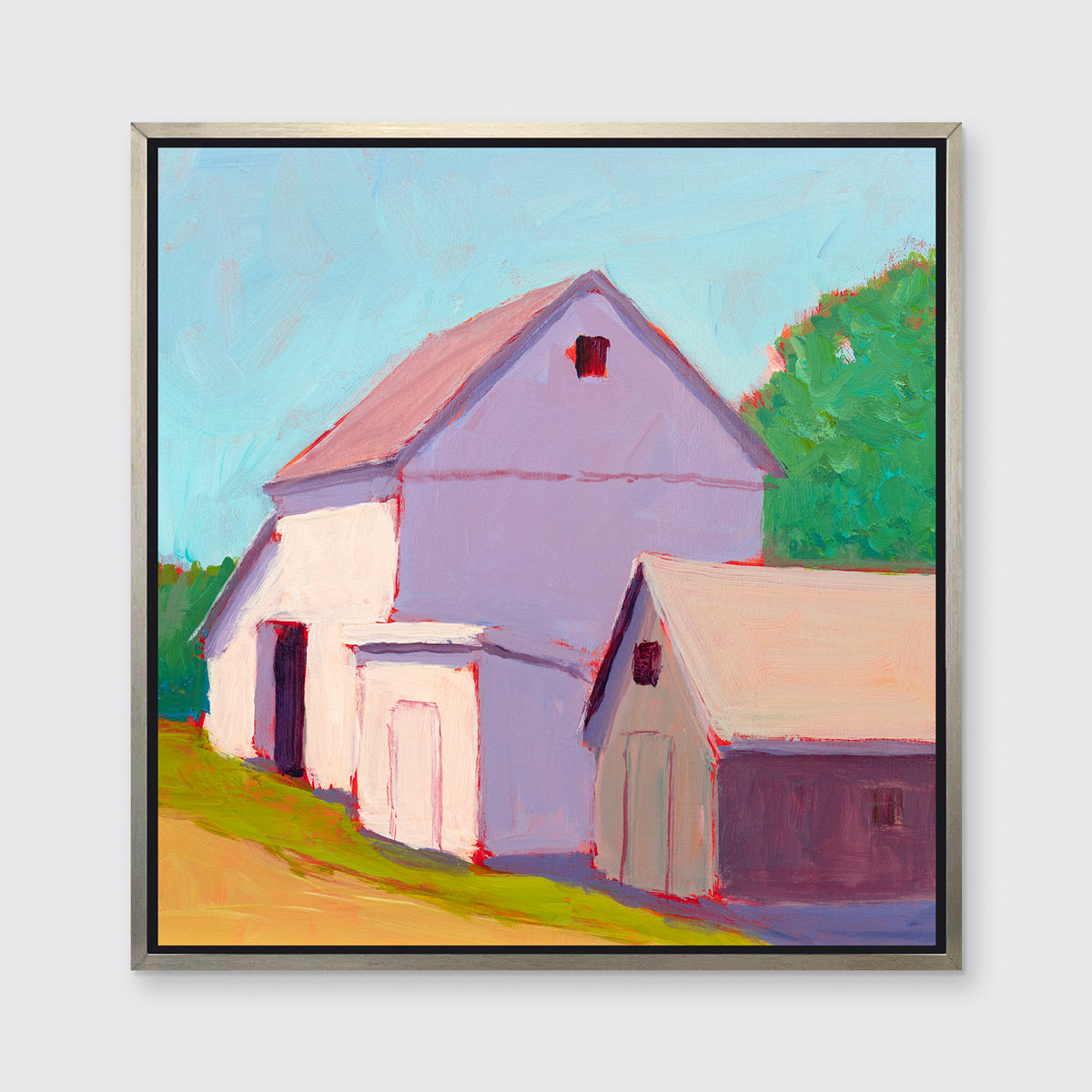 A print of a ivory barn, a smaller tan barn, a blue sky, green trees and a tan road in a silver floater frame hangs on a white wall.