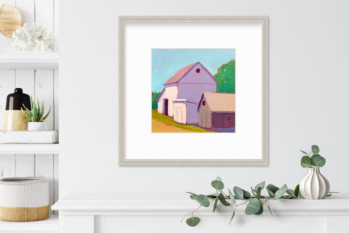 A print of a ivory barn, a smaller tan barn, a blue sky, green trees and a tan road in a silver frame with a mat hangs on a white wall above a white shelf with a small white vase with eucalyptus and next to a built in bookshelf with decorative accessories.
