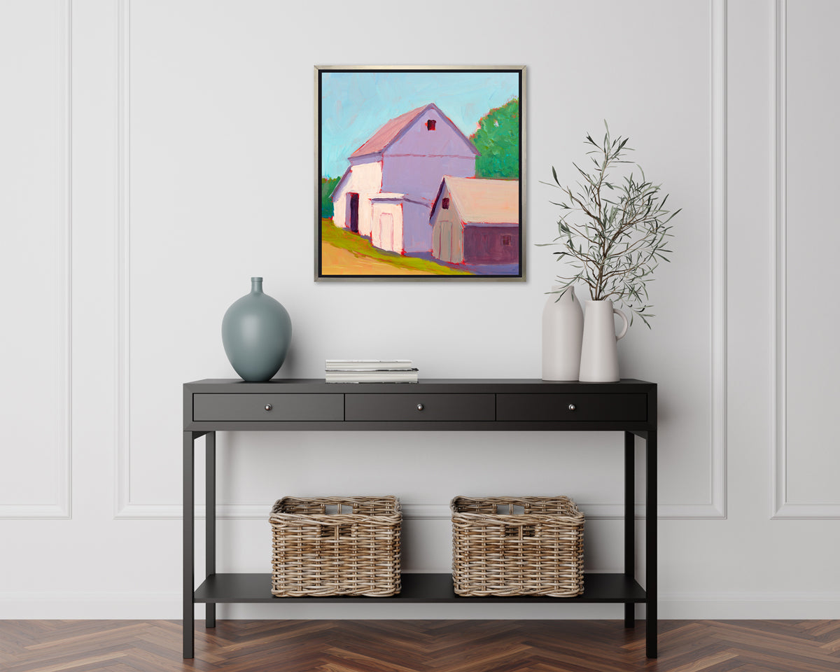 A print of a ivory barn, a smaller tan barn, a blue sky, green trees and a tan road in a silver floater frame hangs on a white wall above a black console table with a muted teal vase, a stack of books, two white vases with greenery and two woven baskets.