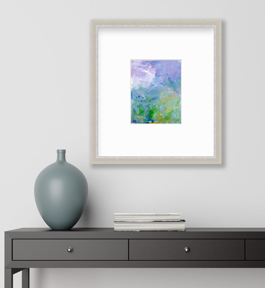 A blue, purple and green abstract print in a silver frame with a mat hangs on a white wall above a black console table with a stack of books and a muted teal vase.