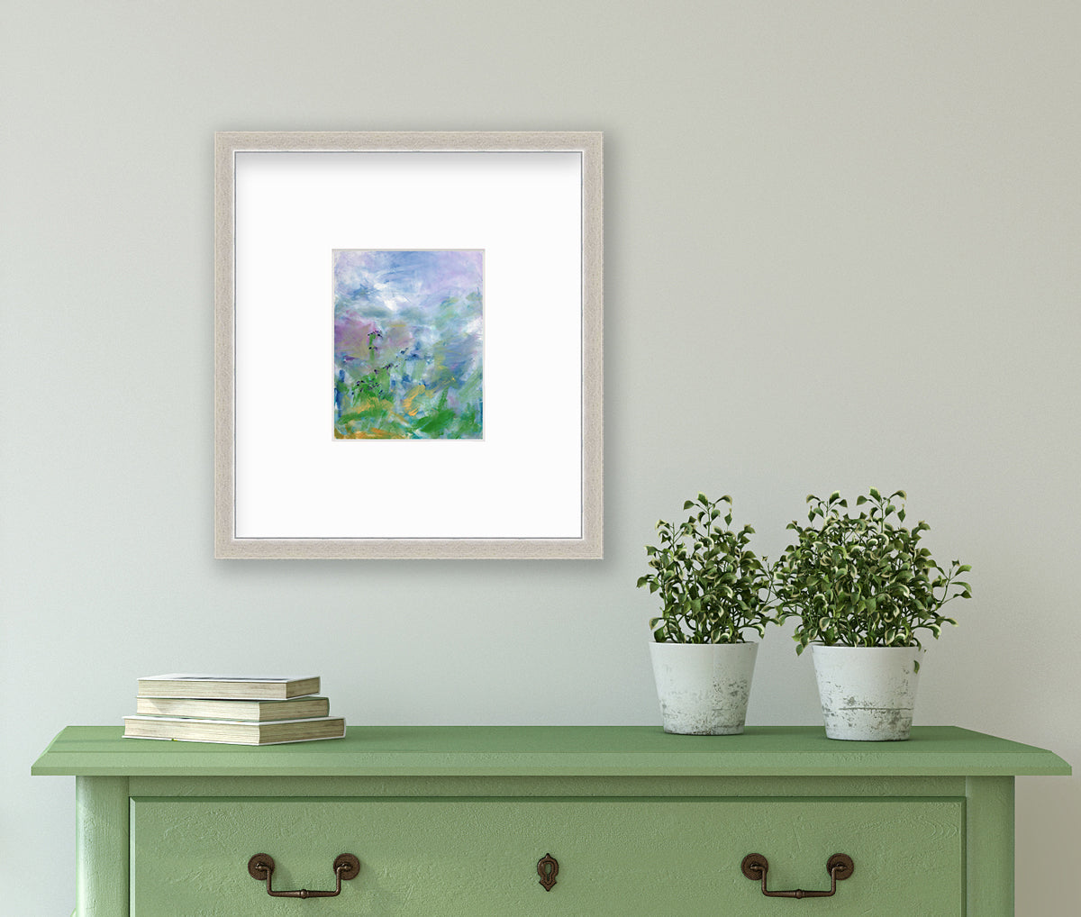 A blue, purple and green abstract print in a silver frame with a mat hangs on a white wall above a green console table with a stack of books and two potted plants.