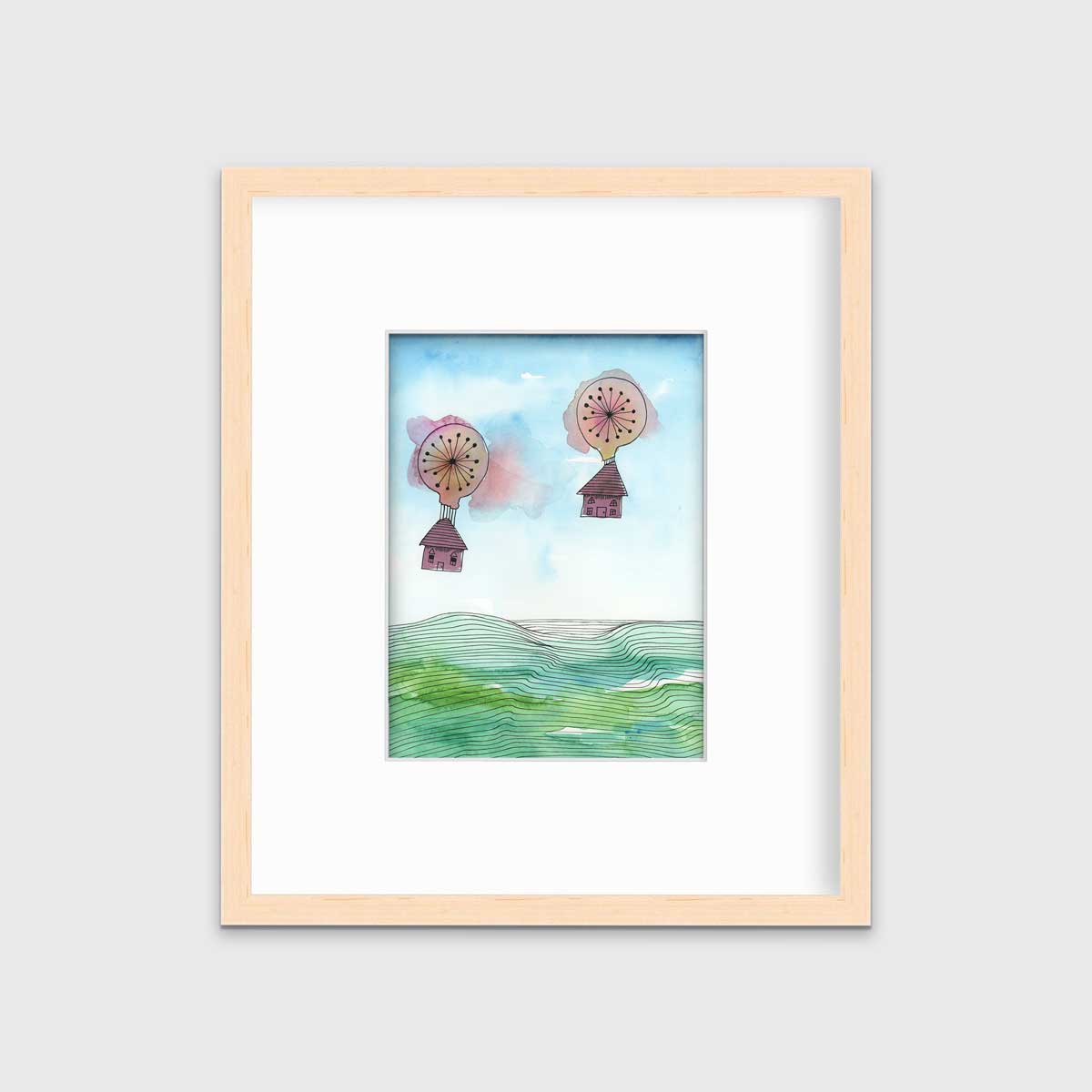 A hot air balloon and house illustration print in a natural wood frame with a mat hangs on a white wall.