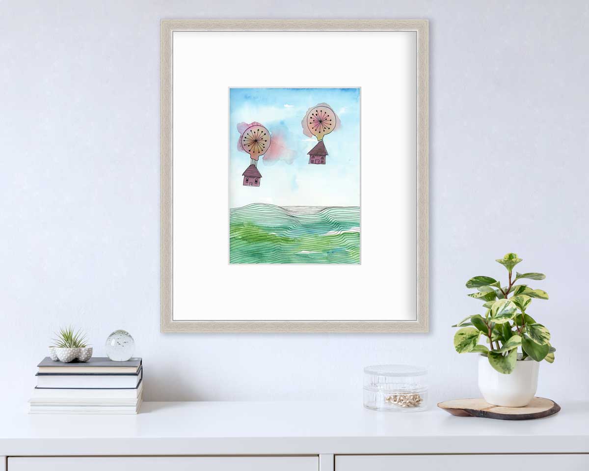 A hot air balloon and house illustration print in a silver frame with mat hangs on a white wall above a white surface with a stack of books, a clear container with gold accessories and a potted plant.