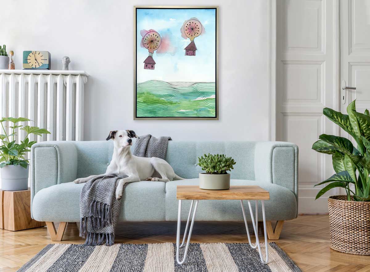 A hot air balloon and house illustration print in a silver floater frame hangs on a white wall above a light blue sofa with a dog and a blue throw blanket, a small side table with a potted plant and two potted plants.