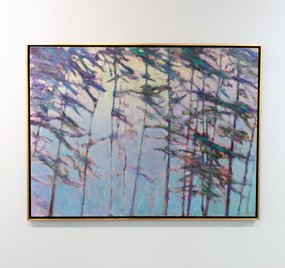 An impressionistic landscape painting by Ken Elliott of trees with blue and purple leaves in a gold floater frame hangs on a white wall in the gallery.