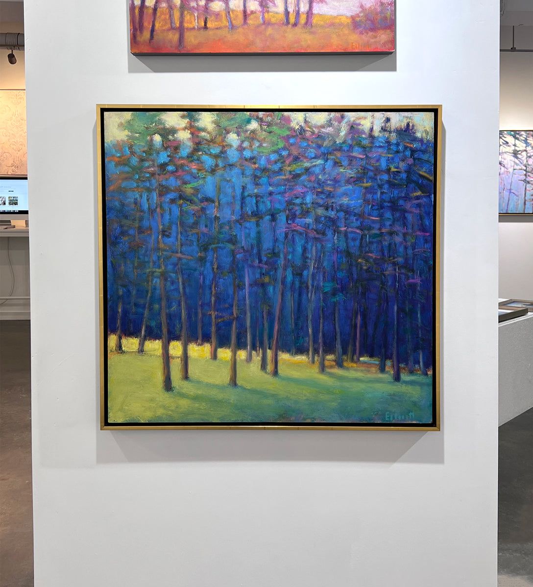 An impressionist painting of a forest with multi-colored leaves and sunlight backlighting the trees from the left in a gold floater frame hangs on a white wall in the gallery.