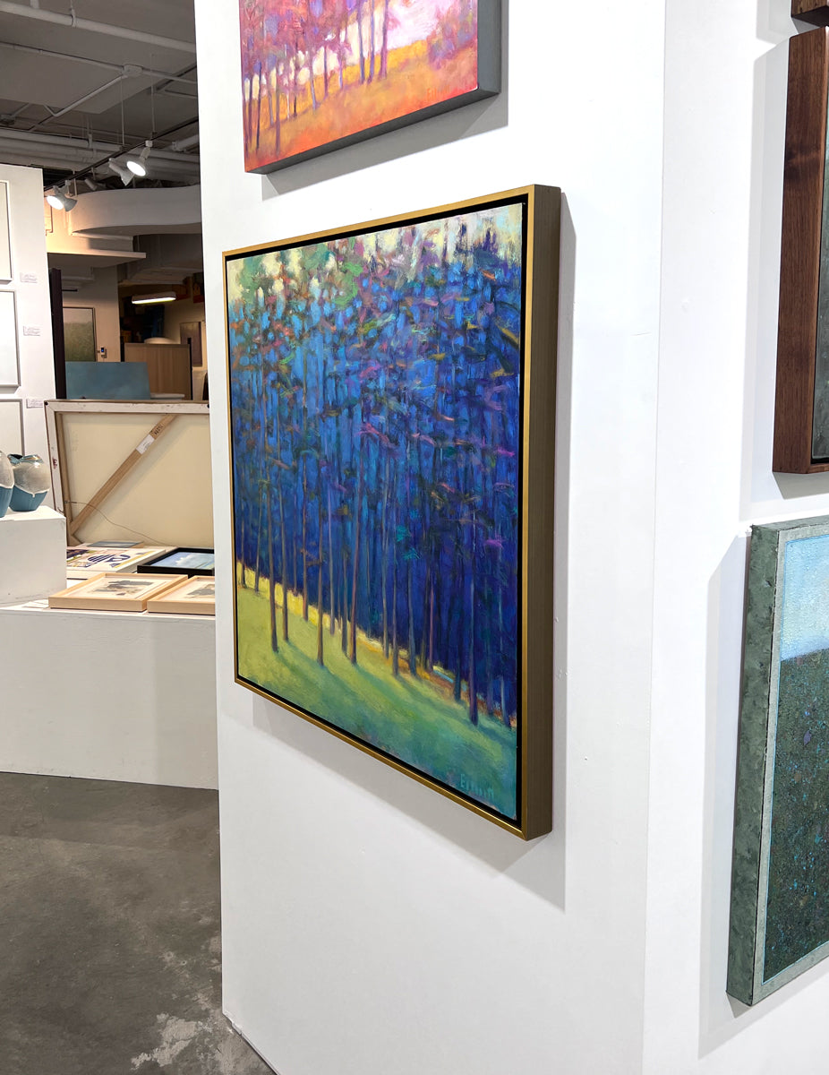An impressionist painting of a forest with multi-colored leaves and sunlight backlighting the trees from the left in a gold floater frame hangs on a white wall in the gallery, viewed from the right. 