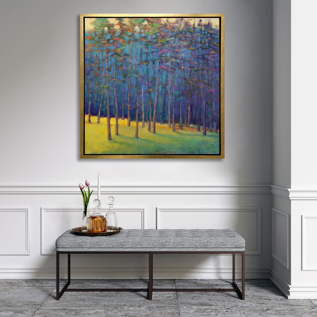 A blue, green and yellow abstract tree landscape print in a gold floater frame hangs on a white wall above a grey tufted bench with a tray with two glass decanters, a white tapered candlestick and a glass vase with pink tulips.