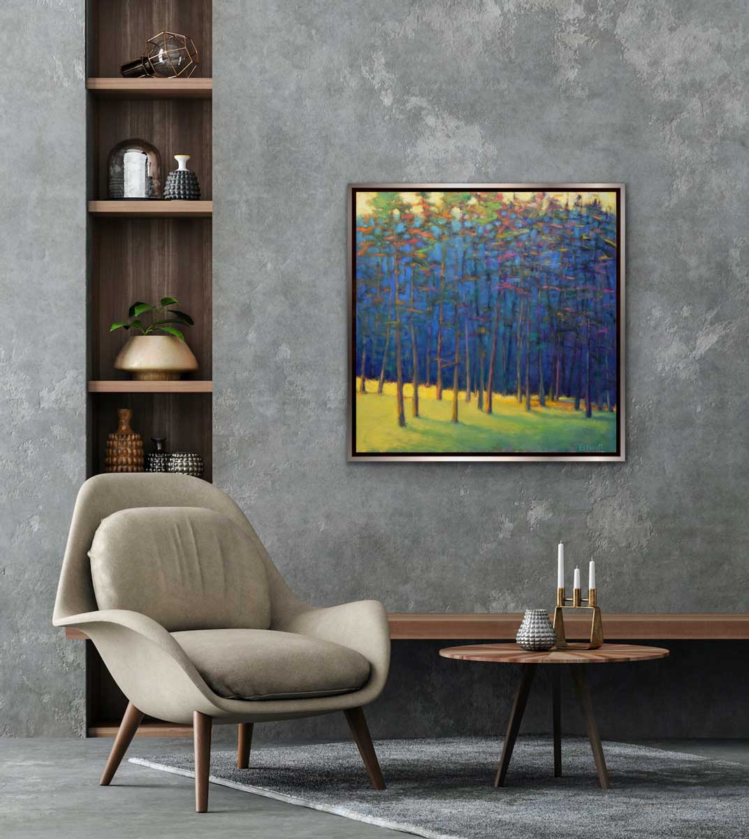 An impressionist painting of a forest with multi-colored leaves and sunlight backlighting the trees from the left hanging on a cement wall above a wooden side table and beige arm chair. The painting hangs to the right of a built in bookshelves with a variety of decor. 