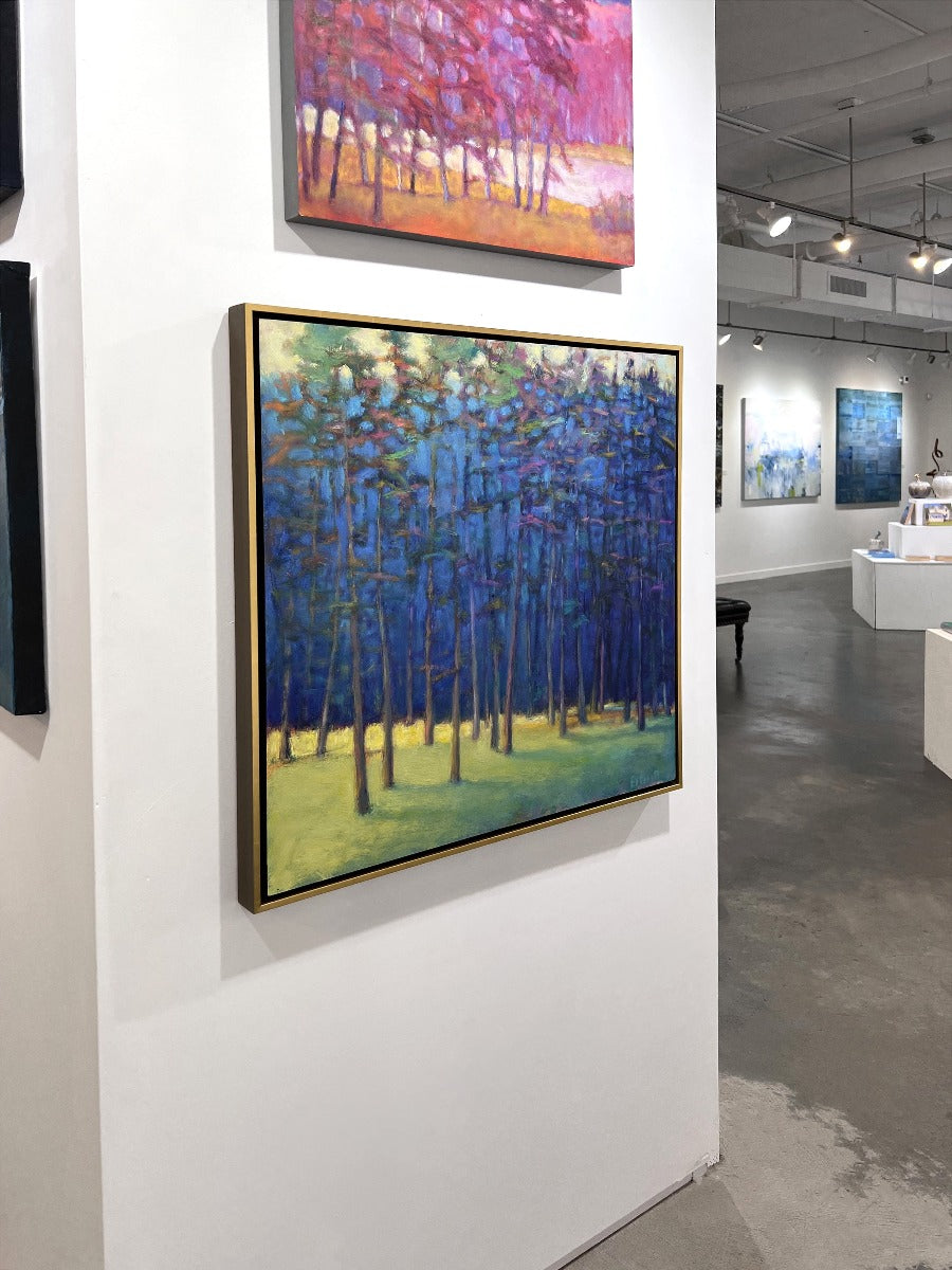 An impressionist painting of a forest with multi-colored leaves and sunlight backlighting the trees from the left in a gold floater frame hangs on a white wall in the gallery, viewed from the left. 