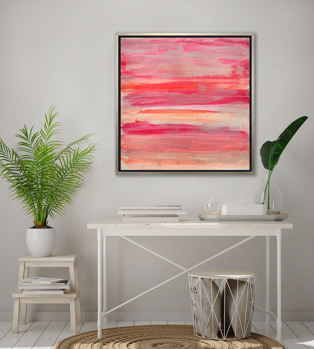 A pink, silver and orange abstract print in a silver floater frame hangs on a white wall above a white desk with a stack of books, a tray with a white box, a clear sphere paperweight and a glass vase with a green leaf stem, a small step stool with a potted plant and a wire basket with pillows.