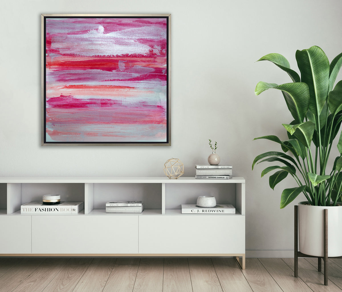 A pink, white, silver and red abstract print in a silver floater frame hangs on a white wall above a white console cabinet with stacks of books, a gold wire sphere and a potted floor plant.